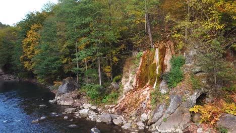 Aerial-shot-of-a-small-waterfall-flowing-down-a-rocky-cliff,-surrounded-by-autumn-trees