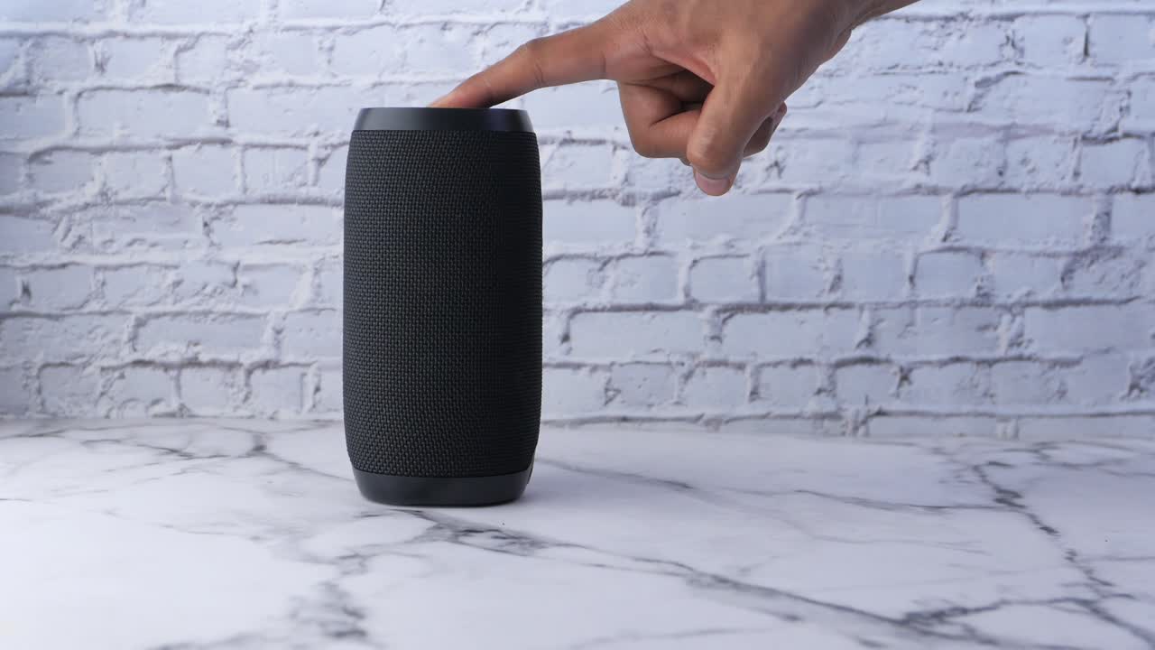mano sosteniendo y tocando un altavoz bluetooth portátil negro en una mesa de mármol frente a una pared de ladrillo blanco