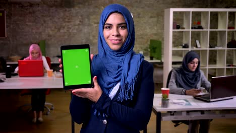 happy smile of arabian woman who is showing green screen on her tablet and standing in modern office, arab women on background, working vibes