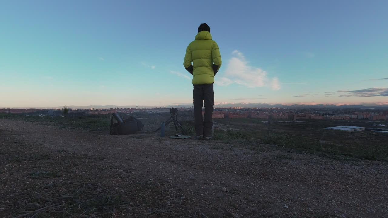 Vista da dietro di un giovane fotografo in piedi con un piumino giallo che guarda lo skyline di Madrid all'alba.