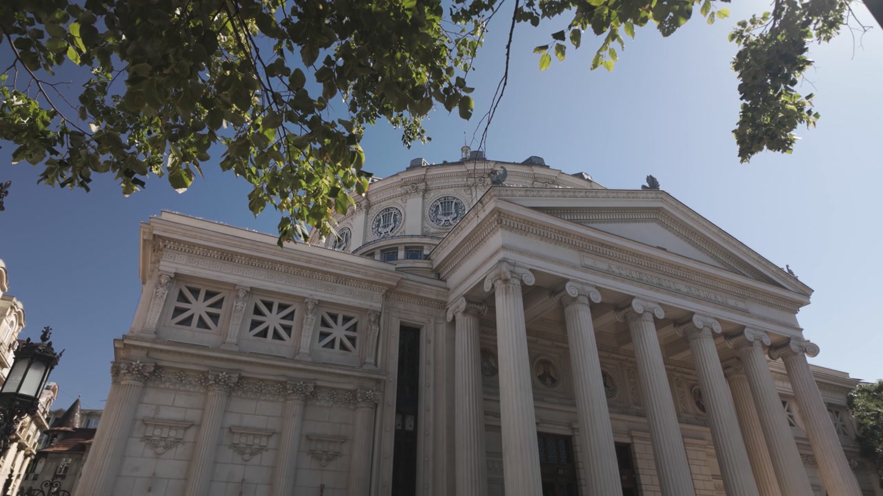 Exterior view of the Romanian Athenaeum in Bucharest, naturally framed by green tree leaves, blending classical architecture with nature.