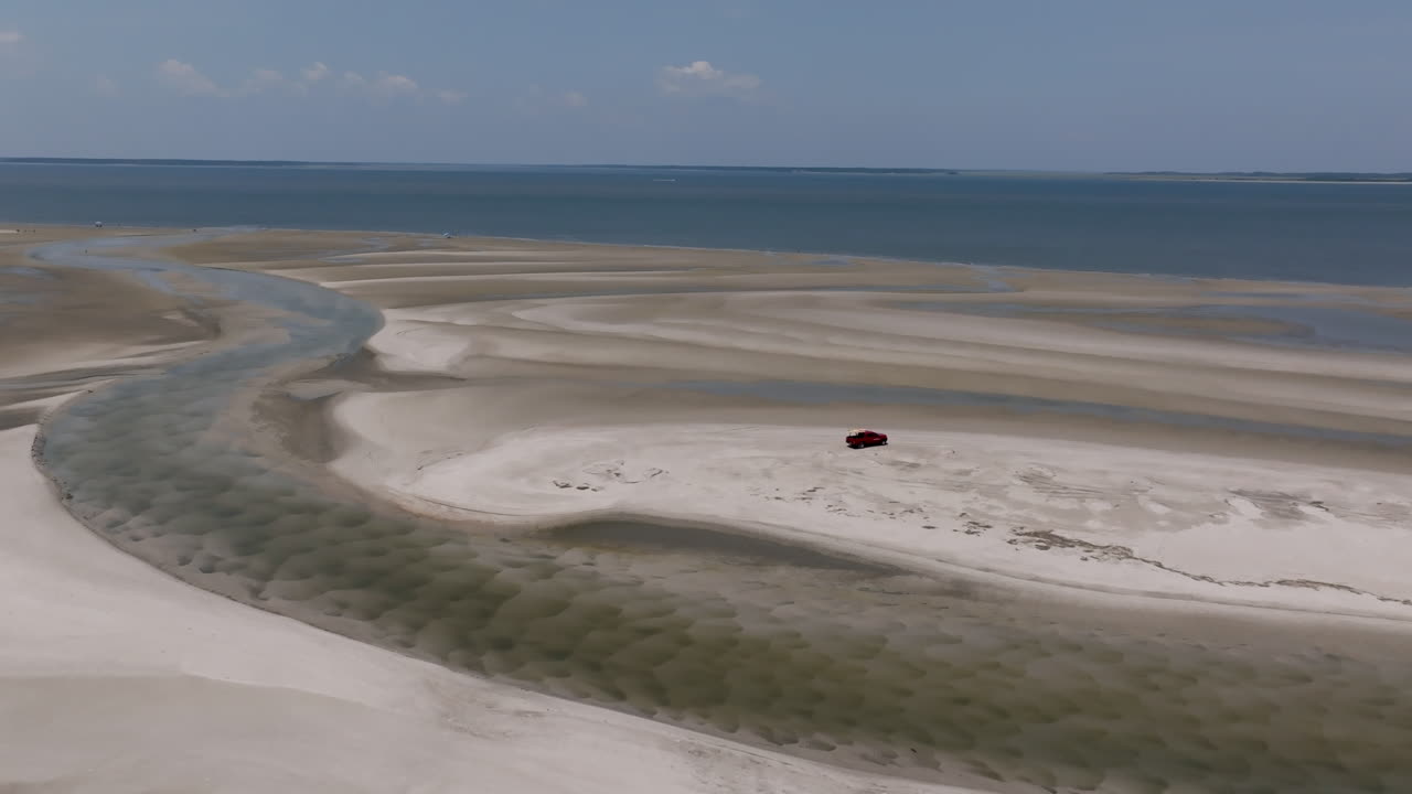 Aerial view of curving tidal sandbars meeting calm blue water, with a lone red SUV parked on a pristine patch of white sand
