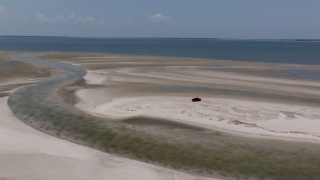 aerial view of curving tidal sandbars meeting calm blue water, with a lone red suv parked on a pristine patch of white sand