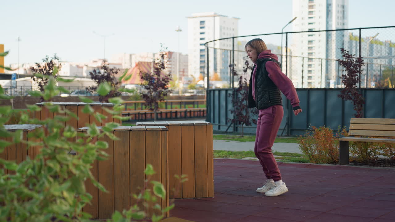 Outdoor trainer bouncing lightly on toes beside wooden park seat in urban park, shifting weight in casual warmup under city skyline during autumn fitness routine, showcasing balance focus and agility