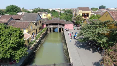 Drohnen-Luftaufnahme-In-Vietnam,-Die-über-Den-Braunen-Flusskanal-Von-Hoi-An-In-Der-Stadt-Fliegt,-Kleine-Backsteinhäuser,-Japanische-Rosa-Alte-Brücke-An-Einem-Sonnigen-Tag