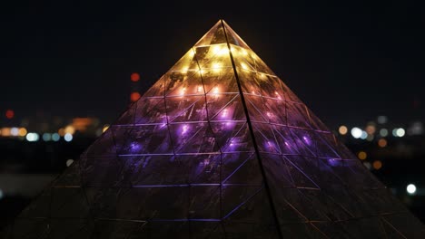 mesmerizing illuminated pyramid at night: a stunning display of colors and reflections captured in vibrant detail with twinkling city lights in the background