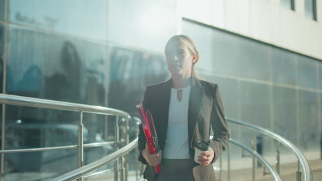 close up of banker in black suit holding file folder and coffee cup while walking along path with blurred background of parked cars and urban buildings during bright morning commute to office