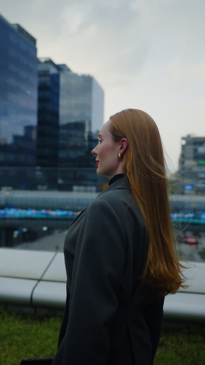 Confident businesswoman with long hair in blazer walks slowly on rooftop terrace of Pangyo office complex with glass buildings
