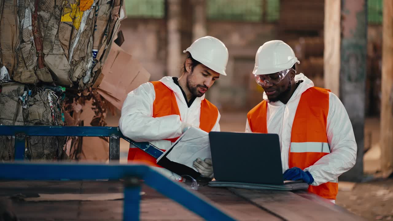 A brunette man with a beard in a white protective uniform in an orange vest talks to his colleague a man with Black skin who works at a laptop while at a large waste processing and sorting plant
