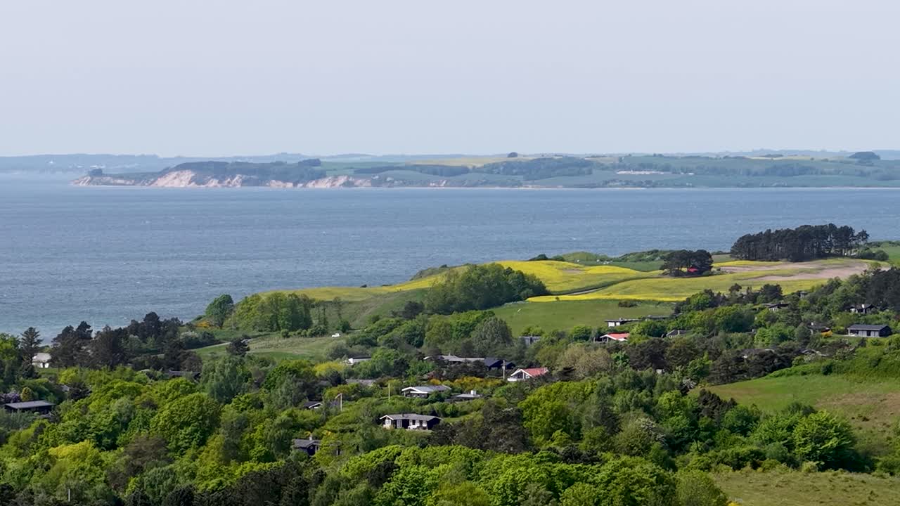 Aerial footage showcases a coastal farmland scene in Denmark. A lush green field and adjacent tilled land are separated by a straight dirt path, all beside calm blue water under an open sky