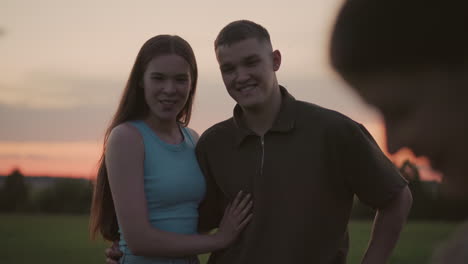 close blur view of hand holding blue pen writing on paper while young woman and man smile watching against dusky sky and field landscape