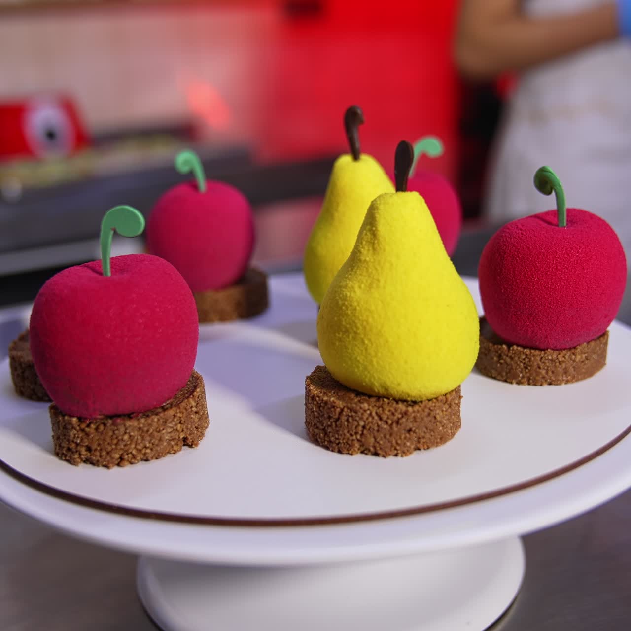 Lovely cakes in the shape of pears and apples on the stand. Confectioner's hands take the pastry and put it on the plate. Close up