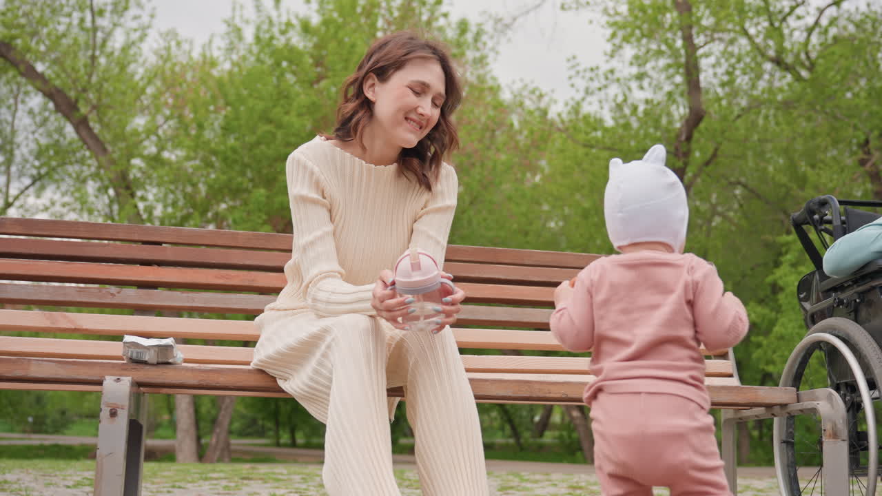 Hvid mor sidder på bænk i parken og opmuntrer tumling i pink jumpsuit nær klapvogn med varmt støttende smil og grønne træer, træbænk, afslappet outfit, hyggestund og blid vejledning