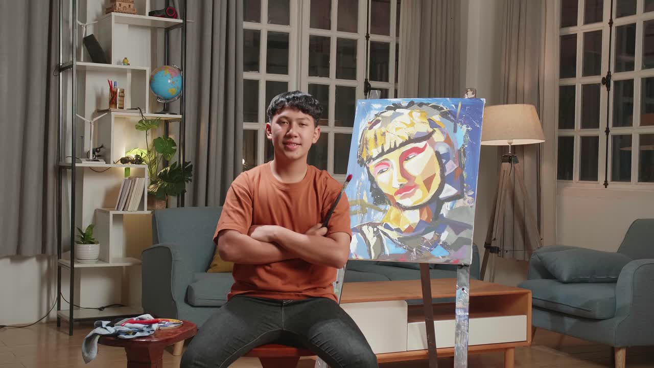 Asian Artist Boy Sitting On Round Wooden Chair Without Backrest Holding Paintbrush Mixed Colour, Crossing His Arms, And Smiling After Finish Painting A Girl On The Canvas