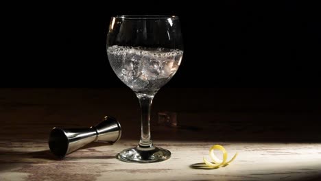 person pouring ice cubes into glass of gin tonic with lemon and jigger on table in dark room