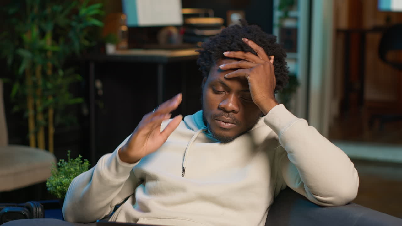 Self employed man suffering from pain and high fever sitting on sofa, trying to work and finish his tasks. Black male feelancer suffering from illness and feeling sick, using laptop. Camera B.