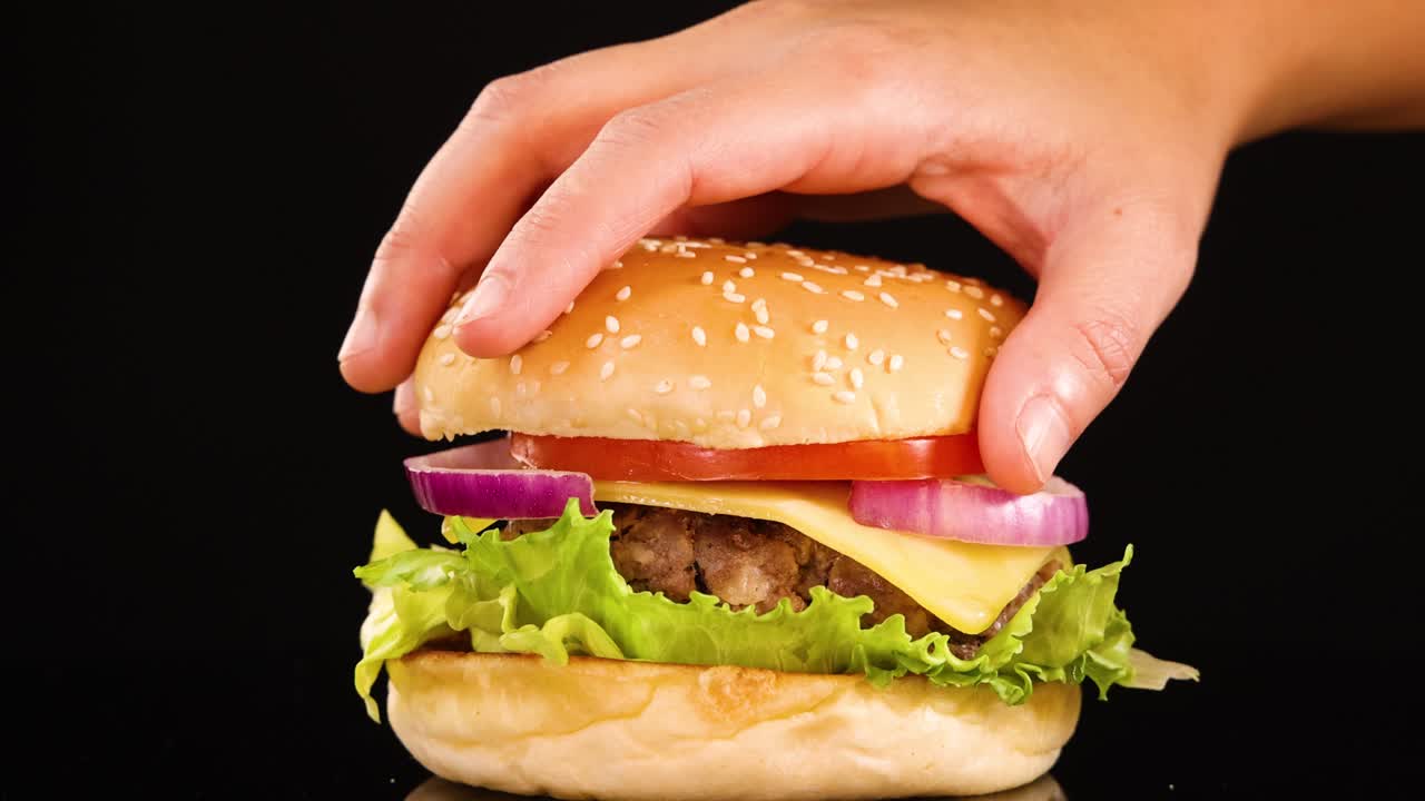 A hand carefully places a sesame seed bun atop a beef burger with lettuce, tomato, onion, and cheese against a clean black background using bright studio lighting
