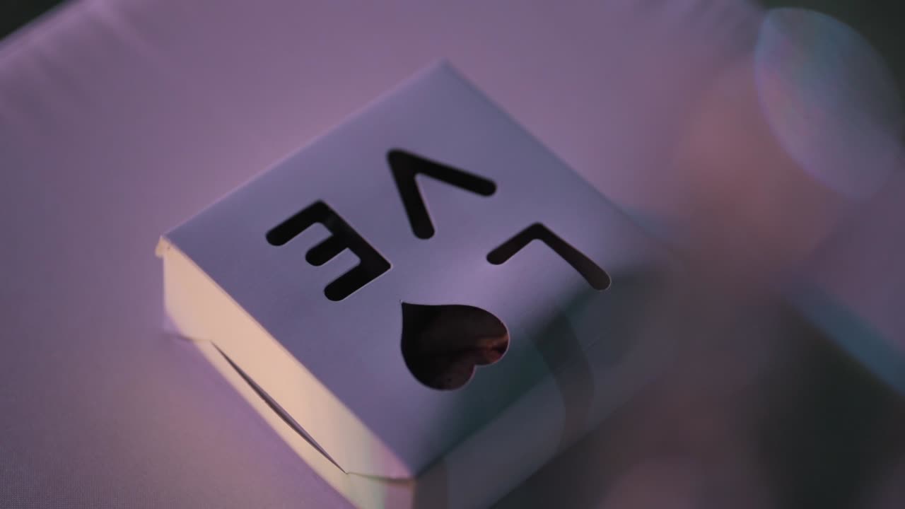 Allestimento per cerimonia nuziale con piccola scatola regalo bianca con dettaglio a forma di cuore posizionata sul sedile di una sedia sotto una morbida illuminazione ambientale con profondità di campo ridotta, ripresa in slow motion con rivelazione.