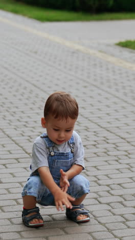 cute baby boy in jeans romper squats on the paved road. interested kid touches the tiles on the ground. vertical video