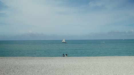 people relaxing on a pebble beach with a sailboat in the distance