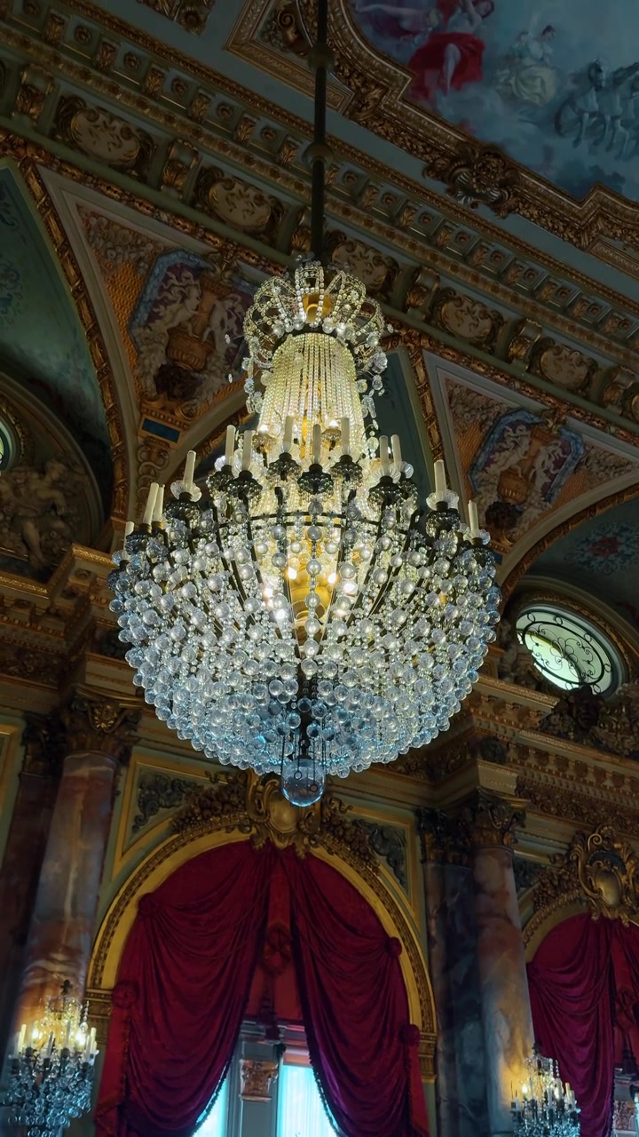 Interno opulento di un palazzo con grande lampadario di cristallo e drappeggi rossi. Vista verticale di una stanza lussuosa con colonne di marmo, dettagli dorati e un ampio lampadario di cristallo sospeso.