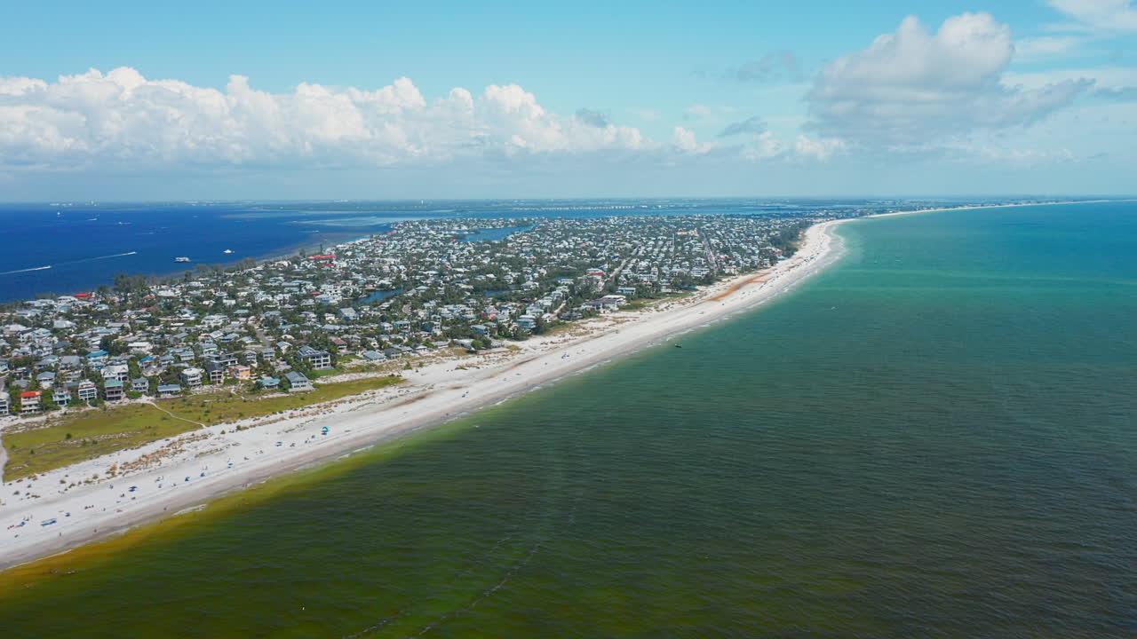 The shoreline of Anna Maria Island stretches along the turquoise ocean, where white sandy beaches meet a bustling seaside community beneath a partly cloudy sky