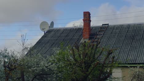 Satellite dishes and a red brick chimney on a tin roof house, surrounded by spring trees under a cloudy sky. Daugavpils, Latvia (Latgale)