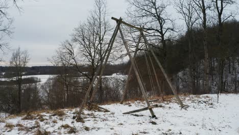 Alte-Holzschaukel-In-Der-Nähe-Eines-Hügels-Bewegt-Sich-Bei-Wind,-Bewölktem-Und-Verschneitem-Wintertag