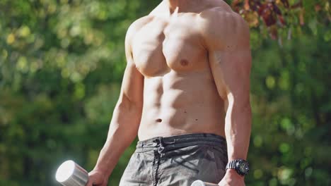 strong body of a bodybuilder exercising outdoors. muscular young man picks up dumbbells in his hands among nature.