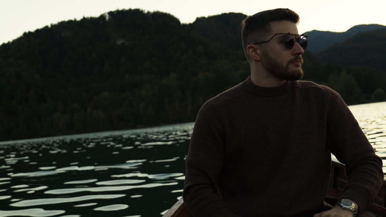 A man rows a wooden boat during sunset, surrounded by moody autumn colors. Peaceful outdoor scene with warm evening light over the water