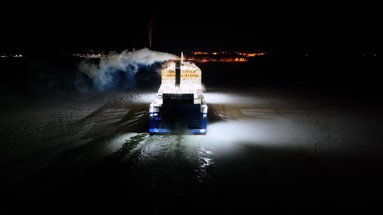 Finnlines hybrid roro ship FINNSIRIUS moving through ice covered sea at night. Harbour lights ahead. Static low altitude aerial stern view.