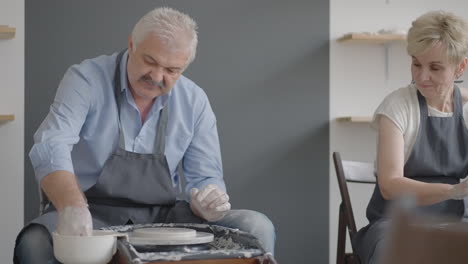 medium shot of middle aged ceramic artist teaching group elderly caucasian woman and senior man how to wedge clay sitting at desk in art studio. people enjoying talking at work