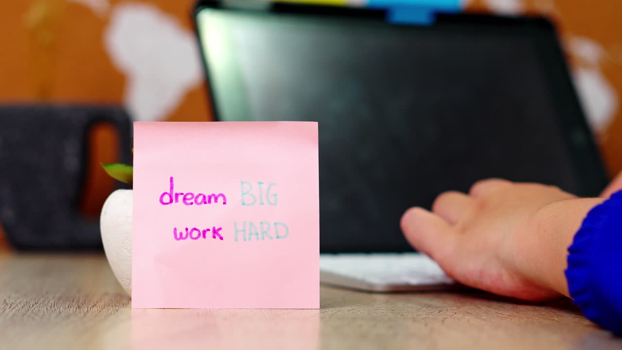 Close-up of Hand placing sticky note with "dream BIG work HARD" on desk front of plant