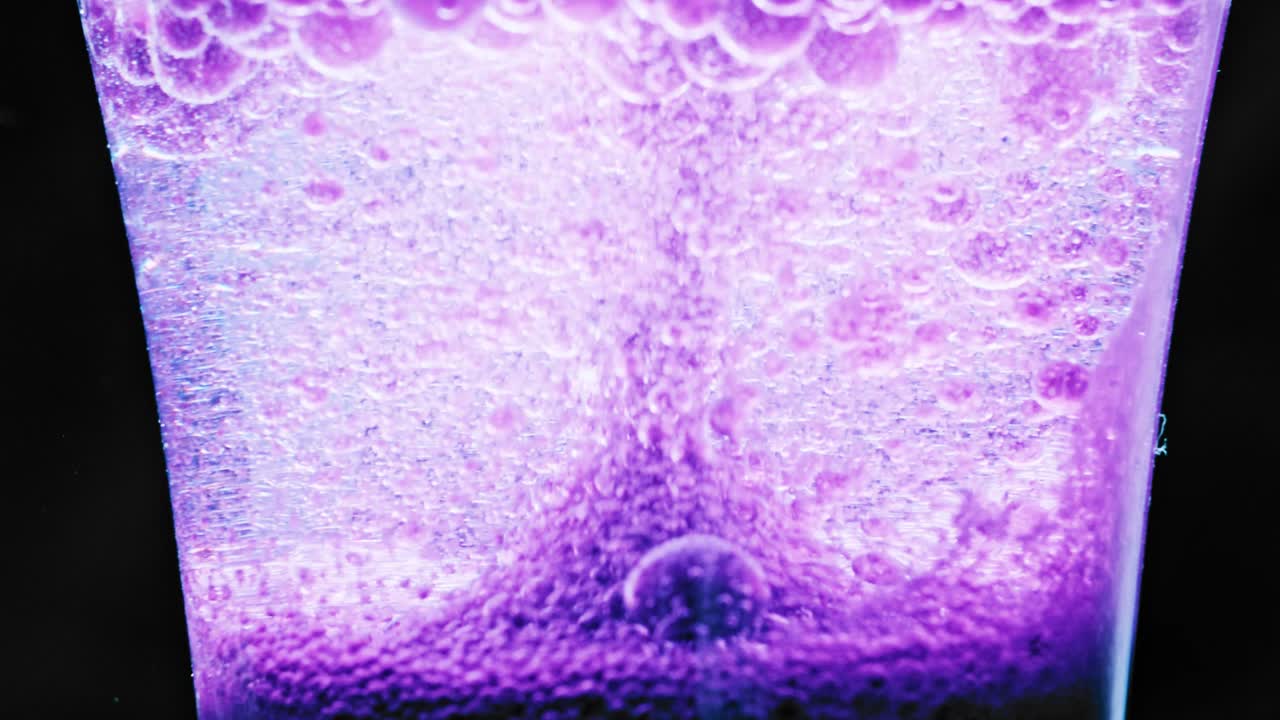 макросъемка искрящихся фиолетовых пузырьков, движущихся вверх в бокале на светлом фоне, и нескольких больших пузырьков, стекающих вниз