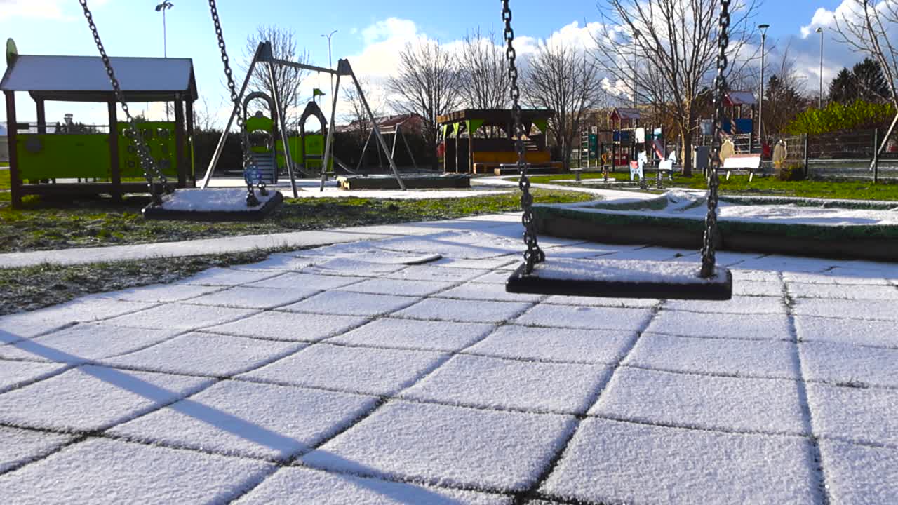 Slow motion video of two playground swings swinging back and fourth that are covered in fresh first white snow during a sunyn day with blue sky and green grass in the background covered in snow