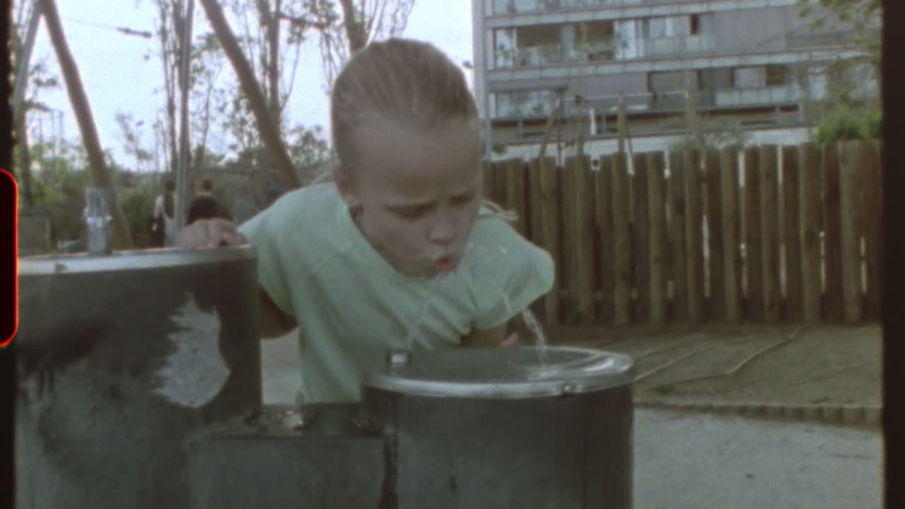 Filmed on super 8 film - youthful girl drinking water from fountain in a playground during sunny summer day, capturing nostalgic vintage moment of carefree childhood in urban playground