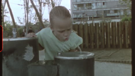 filmed on super 8 film - youthful girl drinking water from fountain in a playground during sunny summer day, capturing nostalgic vintage moment of carefree childhood in urban playground