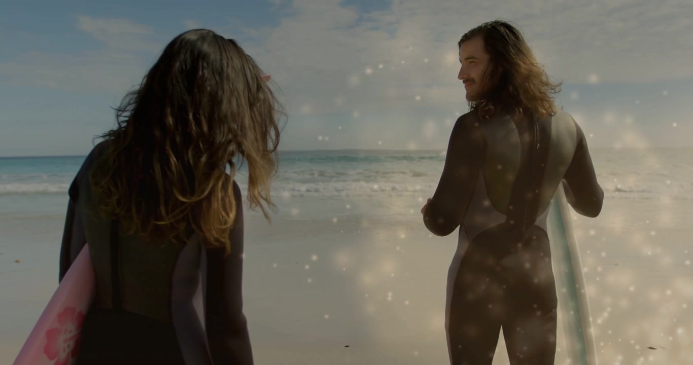 Kaksi surffaajaa kävelee kohti merta, toinen säätää hihnaa, hymyillen, valon heijastus kimaltelee surffauksen yllä.