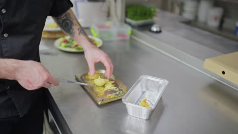 close-up view of a confident male cook in a black uniform transfers ready-made dishes from a baking dish into a packed box for home delivery of dishes