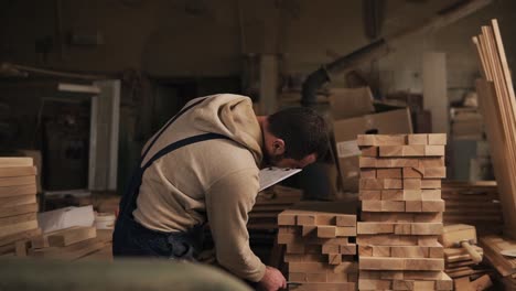 Un-Hombre-En-Un-Taller-De-Carpintería-Con-Una-Tableta-Y-Un-Bolígrafo-Cuenta-El-Número-De-Bloques-De-Madera.-Ensamblador-Está-En-La-Ropa-De-Trabajo.-Vista-Frontal