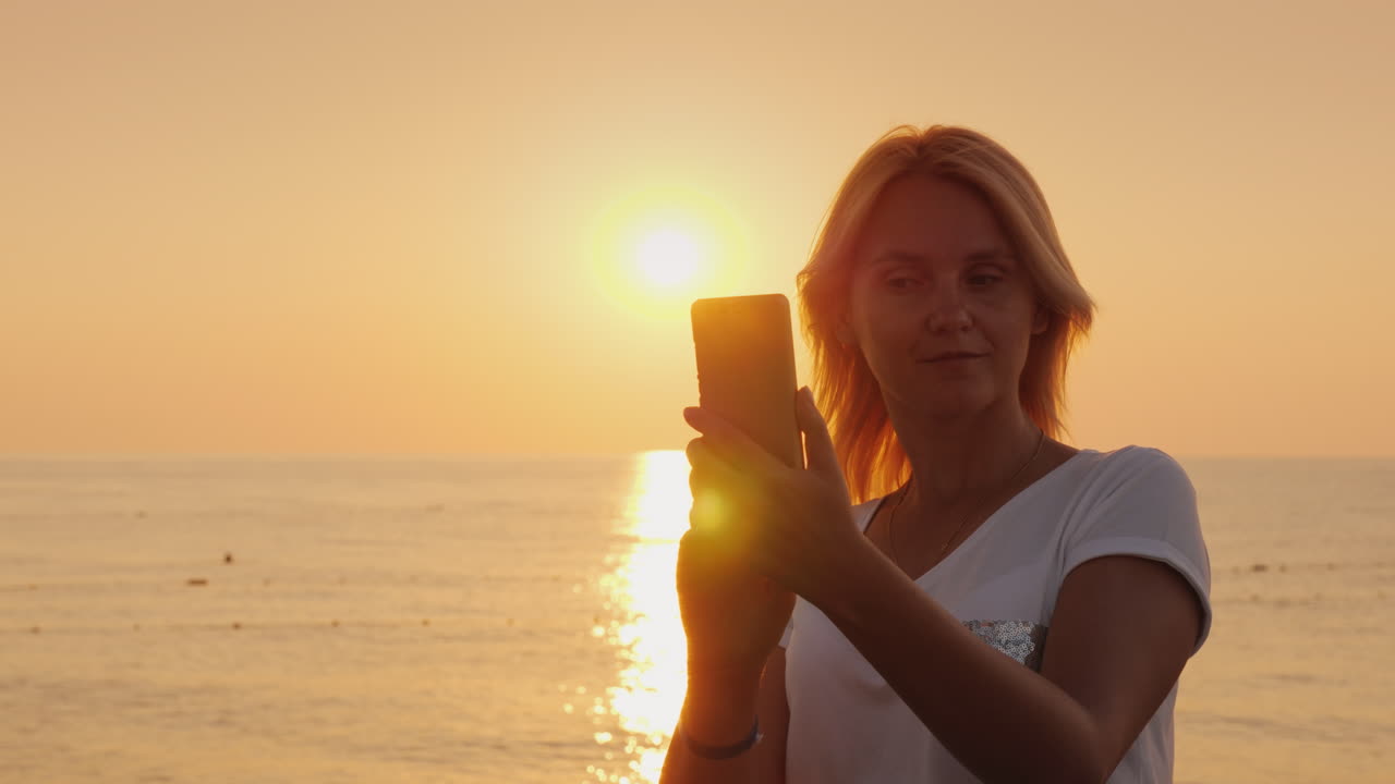 una joven rubia está fotografiando en un smartphone un amanecer rosa junto a la vista del mar desde atrás
