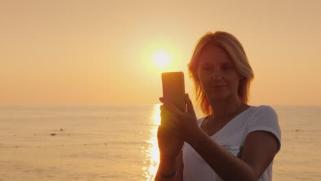 Eine-Junge-Blonde-Frau-Fotografiert-Auf-Einem-Smartphone-Eine-Rosa-Morgendämmerung-Am-Meerblick-Von-Hinten