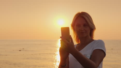 a young blonde woman is photographing on a smartphone a pink dawn by the sea view from the back