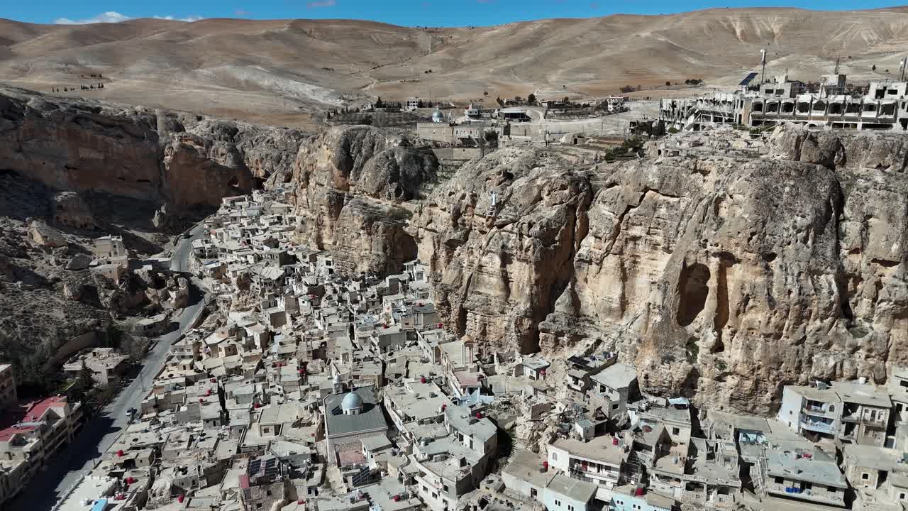 Wide aerial panorama of Ma'loula, Syria, captured from the surrounding arid slopes, showing the ancient village spread along the base of massive desert cliffs