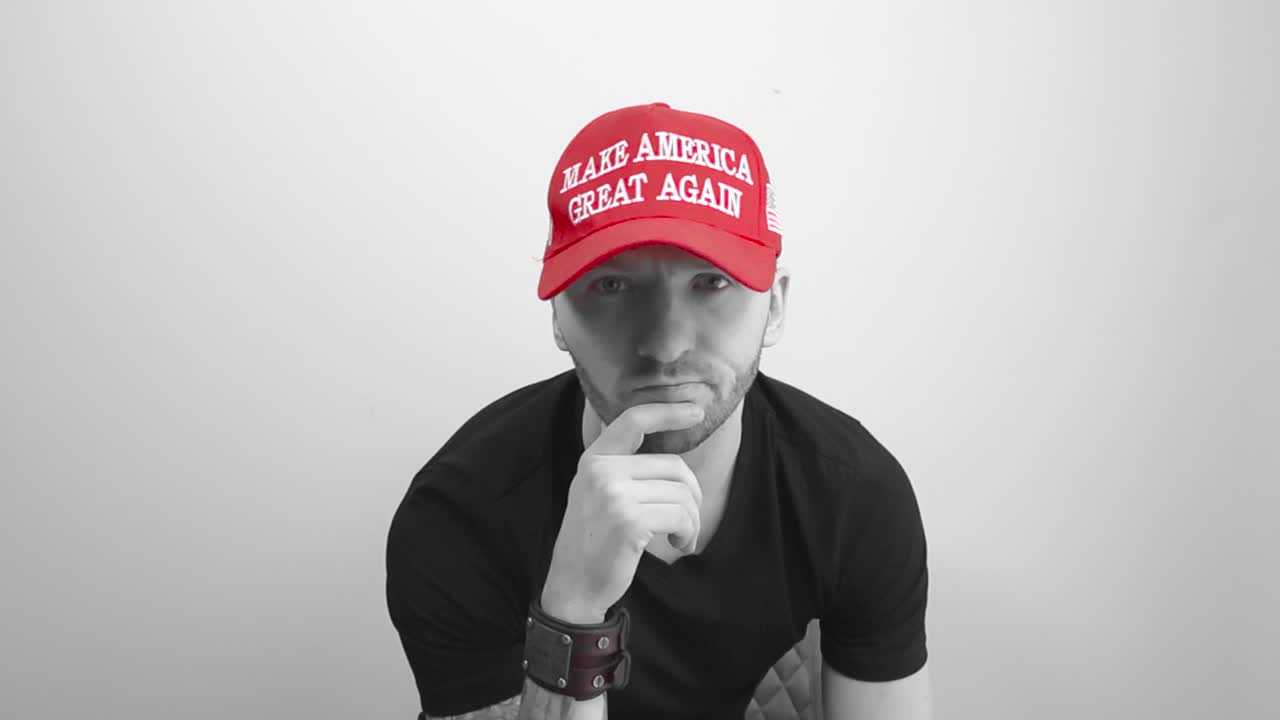 Grayscale footage of a tattooed man with a red colored MAGA Make America Great Again hat sitting with his hand on his jaw and looking forward while shaking his head showing unhappy emotion. man sits.