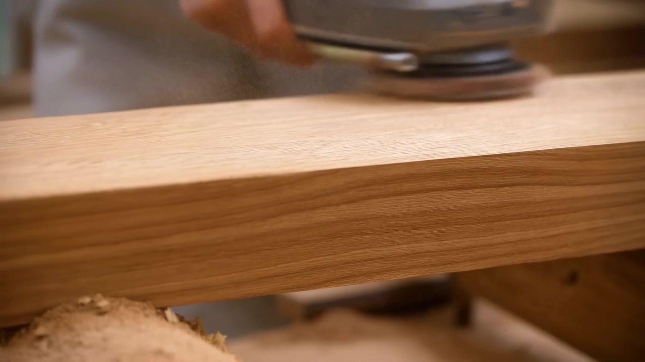 Uma lixadeira orbital manual trabalha em uma prancha de madeira em uma cena de oficina em close-up, suavizando a superfície sob luz natural quente com um fundo desfocado.