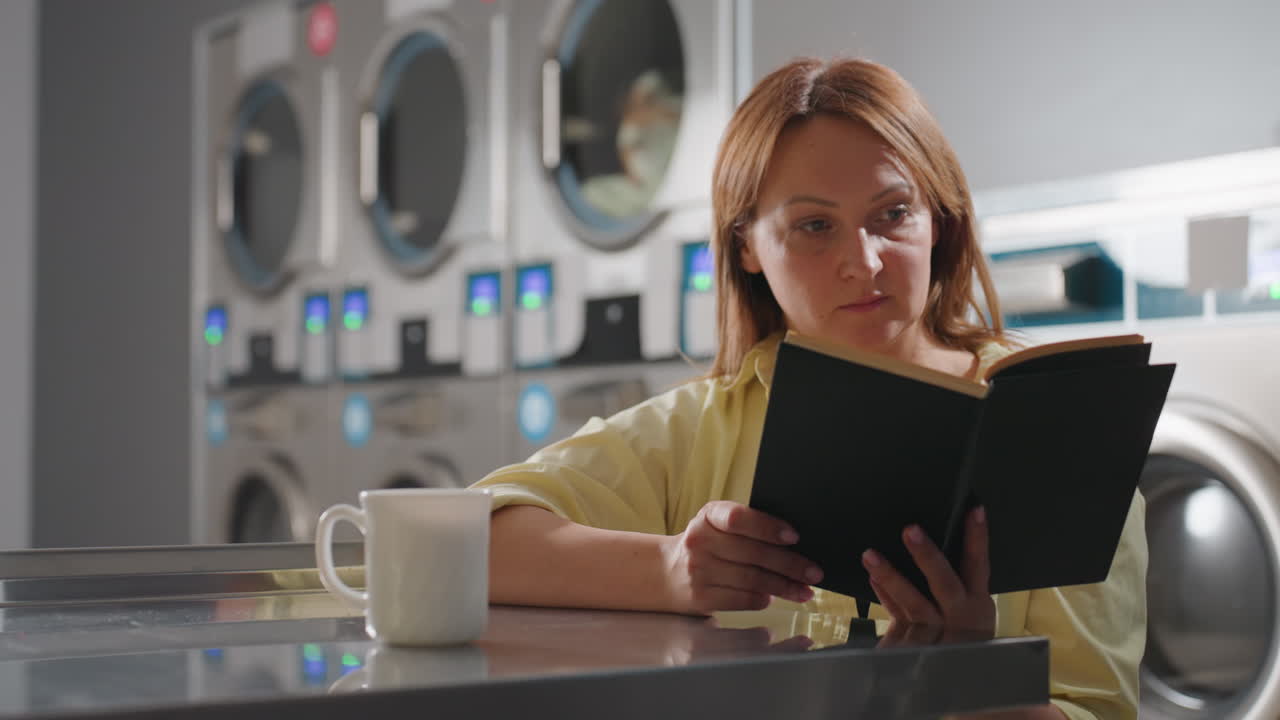 Laundry owner seated at table inside laundromat reading notebook with focus, cup of coffee placed on surface, stainless industrial washers with glowing digital panels in background
