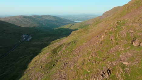 flying close to mountainside illuminated by early morning sunshine. with mountain pass road snaking towards distant lake windermere. summer. kirkstone pass, lake district, cumbria, uk