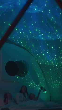 madre con niños se acuesta en una cama blanca y explica las ubicaciones de las estrellas y planetas apuntando a la proyección del cielo en el techo blanco. noche educativa familiar en luz suave