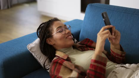 feliz chica afroamericana relajándose en el sofá en casa con un teléfono inteligente
